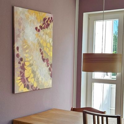 Painting in purple, gold and white colors hangs on a pale purple wall on the head side of a wooden dining table with wooden chairs and a wooden lamp.