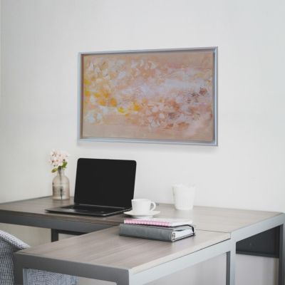 Painting in light pink, beige and white colors hangs on a white wall on the head side of a desk with a laptop and books.