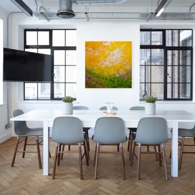 Big square painting in yellow green and gray hanging between two windows on a white wall behind a long white table in a conference room.