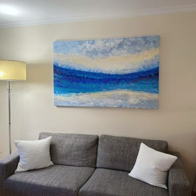 Painting in blue, white and gray hanging on a pale yellow wall behind a gray sofa with white pillows.