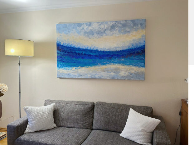 Painting in blue, white and gray hanging on a pale yellow wall behind a gray sofa with white pillows.